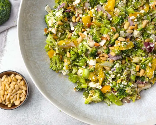 Ein bunter gekochter Brokkoli-Salat mit Pinienkernen, Feta und Orangenstücken auf einem Teller.