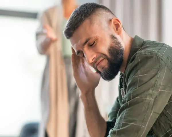 Mężczyzna z ręką na skroni, wyrażający zaangażowanie emocjonalne. W tle niewyraźna postać.