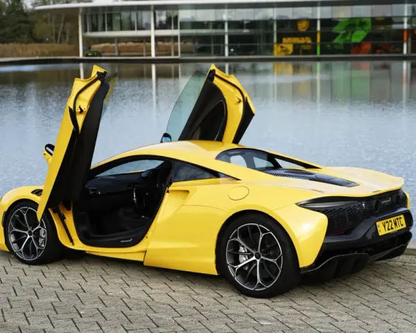 Żółty McLaren Artura z otwartymi drzwiami, stojący nad jeziorem przy nowoczesnym budynku.