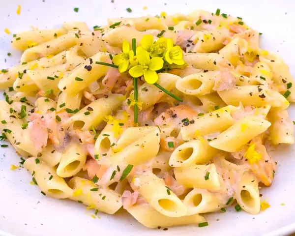 Pasta penne cremosa con trozos de salmón rosado, ralladura de limón y flores amarillas.