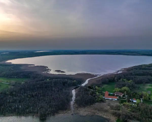 Rozległe jezioro Tuchlin otoczone lasem, z widocznym budynkiem i drogą. Zachód słońca maluje niebo.