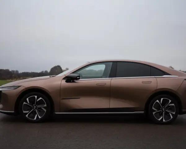 Brązowy samochód elektryczny Mazda6e widziany z boku na tle pochmurnego nieba i krajobrazu.