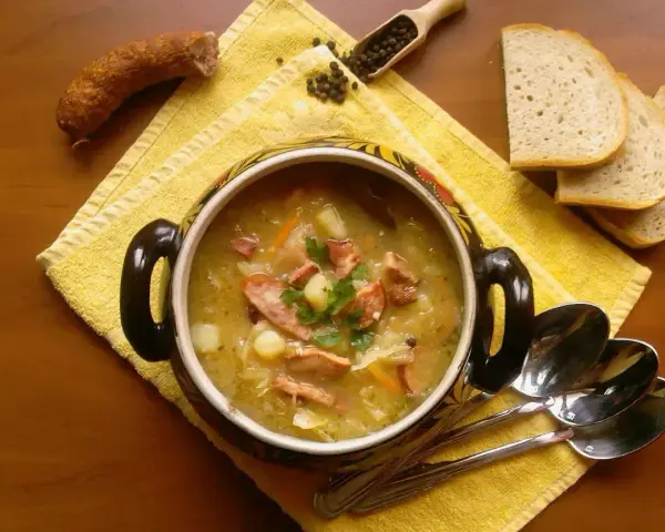 Aromaticzny kapuśniak z kiełbasą i ziemniakami, podany w ozdobnym naczyniu, obok kromek chleba i ziaren pieprzu.