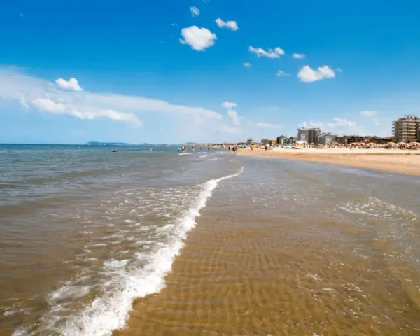 Onda che lambisce la sabbia dorata, con ombrelloni e palazzi sullo sfondo. Un classico scenario di **Emilia Romagna mare**.