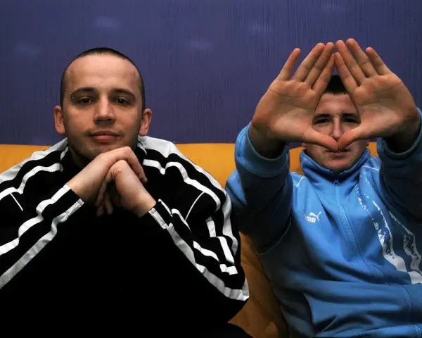 Dwóch mężczyzn w dresach, jeden z rękami złożonymi na brodzie, drugi tworzy rękami kształt serca. Klimat hip hop i rap.