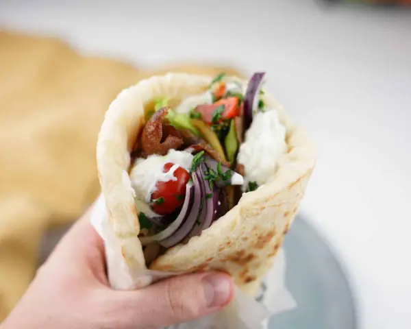 En hand håller en gyros med kött, grönsaker och vit sås. Perfekt för supergod fredagsmat!