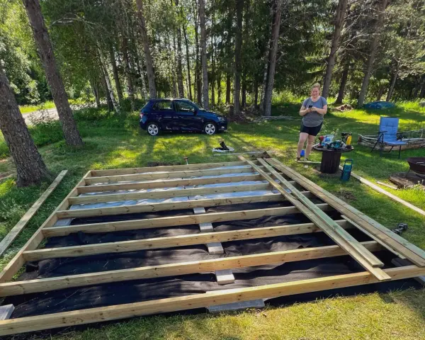 En kvinna förbereder sig för att skruva trall på en nybyggd altan i en lummig trädgård.