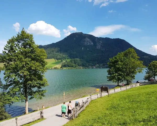 Ein sonniger Tag am bayrischzell see. Menschen spazieren am Ufer entlang, während im Hintergrund ein bewaldeter Berg aufragt.