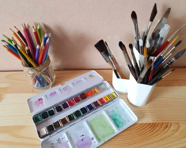 Une palette d'aquarelle ouverte, des crayons de couleur dans un bocal et plusieurs pinceaux indispensables aquarelle dans des pots blancs.