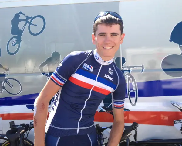 David Gaudu, a French cyclist, smiles in his team's blue, white, and red jersey, with bike graphics on the vehicle behind him.