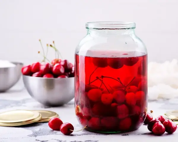 Duży słoik wypełniony rubinowym kompotem z wiśni, obok miska świeżych owoców i cukru.