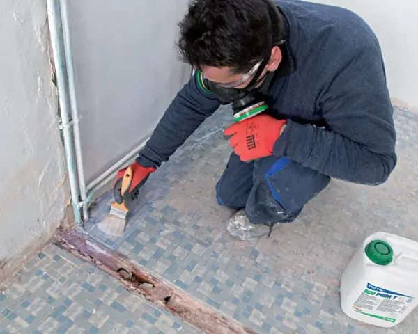 Préparation d'un ragréage sur carrelage ancien. Un ouvrier, masqué et ganté, applique un primaire d'accrochage au pinceau sur le sol carrelé.