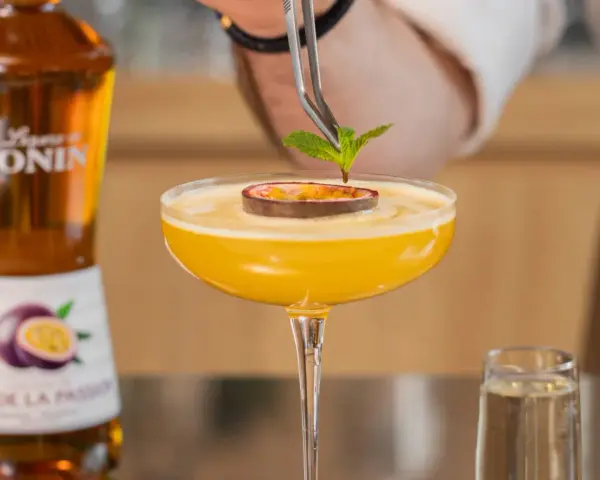 Barman dekoruje drink likierem z marakui, dodając listek mięty. Obok stoi butelka Monin.