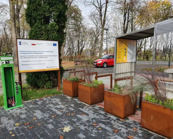 Zielona stacja serwisowa dla rowerów i tablica informacyjna o projekcie VeloMetropolis w Małopolsce. W tle czerwony samochód i jesienne drzewa.