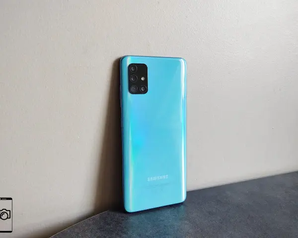 Błękitny Samsung A51 z aparatem w kwadratowej ramce, oparty o jasną ścianę.