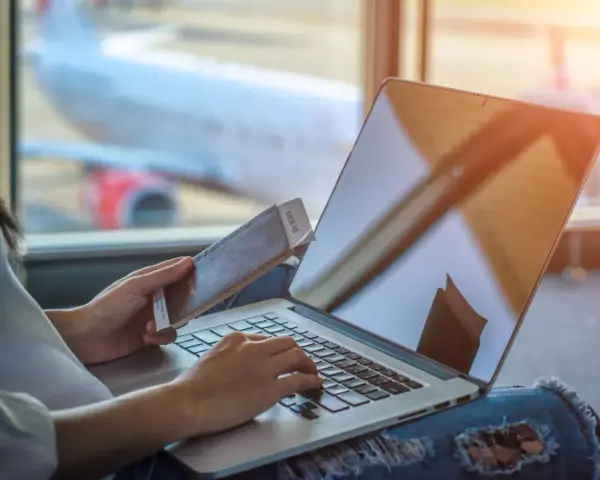 Osoba z paszportem i biletem czeka na lotnisku, sprawdzając na laptopie, ile czasu na odbiór paszportu.
