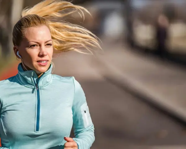 Kvinna med blont hår i hästsvans springer utomhus, lyssnar på musik med hörlurar. Hon är fokuserad på att springa 5 km tid.