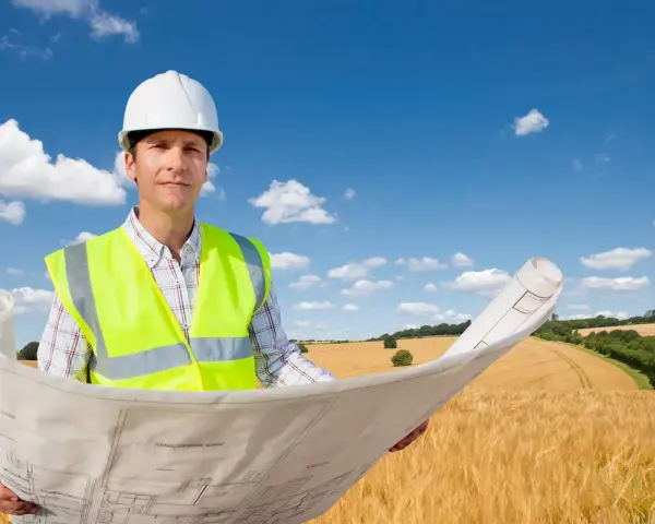 Inżynier w kasku i kamizelce odblaskowej trzyma plany na tle pola. To wizualizacja, jak działka rolno budowlana co to znaczy, czyli połączenie upraw z możliwością zabudowy.