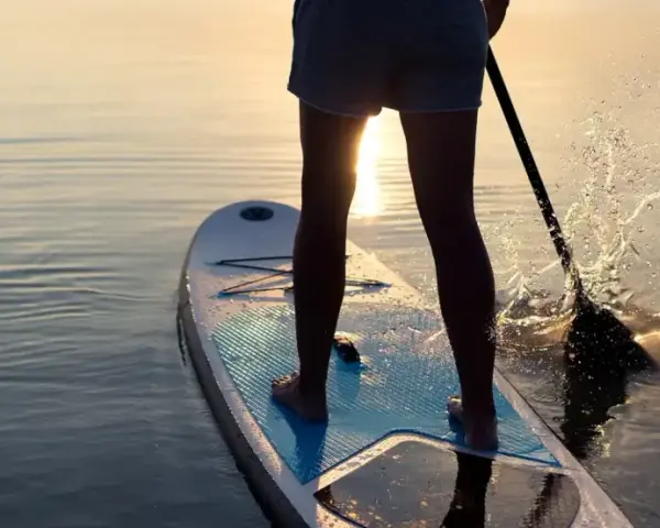 Person lernt SUP auf ruhigem Wasser bei Sonnenuntergang. Das Paddel spritzt Wasser auf.