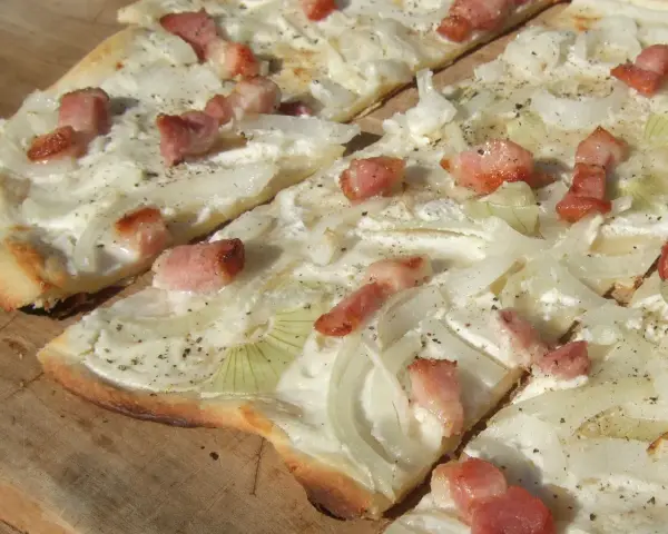 Flammkuchen mit dünnen Zwiebelringen und Speckwürfeln auf Crème fraîche. Ein köstlicher Belag für den knusprigen Teig.