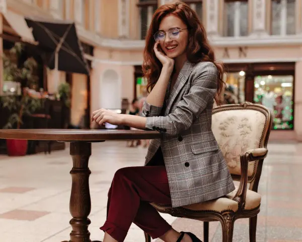 Elegancka kobieta w okularach i marynarce w kratę, prezentująca stylizacje z bordowymi spodniami, siedzi przy stoliku w kawiarni.