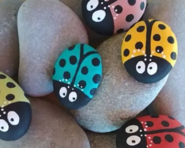Seis piedras pintadas fáciles con mariquitas de colores: roja, amarilla, verde, rosa y turquesa, descansan sobre piedras grises.