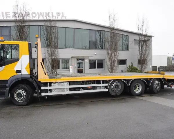 Żółty Iveco Stralis z platformą transportową stoi przed budynkiem.