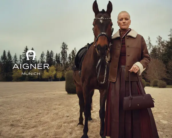 Elegancka kobieta w brązowej kurtce i spódnicy obok konia. Logo AIGNER MUNICH. Aigner co to za marka? Luksusowa moda.