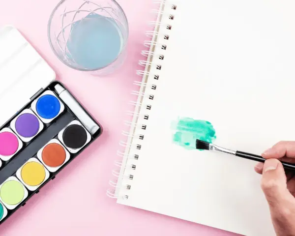 Une main tient un pinceau pour créer une tache verte sur un carnet, à côté d'une palette d'aquarelle et d'un verre d'eau.