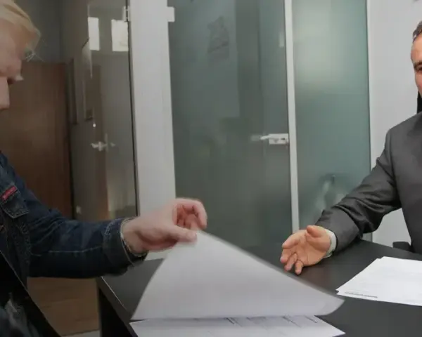 Dwóch mężczyzn przy biurku przegląda dokumenty, jeden w garniturze, drugi w dżinsowej kurtce.