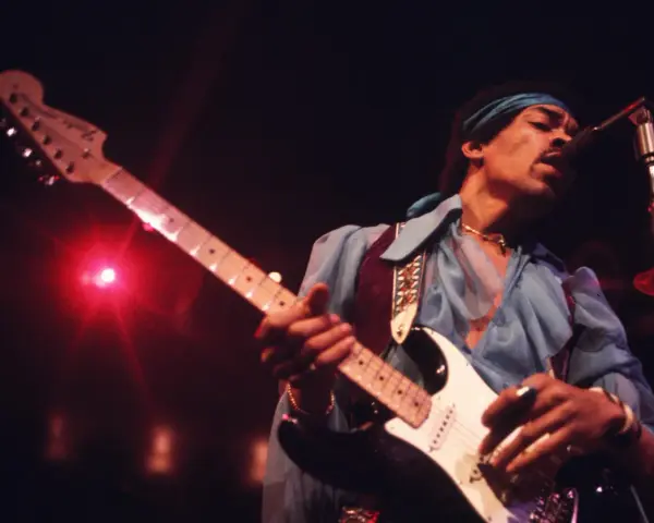 Jimi Hendrix, einer der besten Gitarristen, spielt auf der Bühne seine weiße Fender Stratocaster. Rotes Licht beleuchtet die Szene.