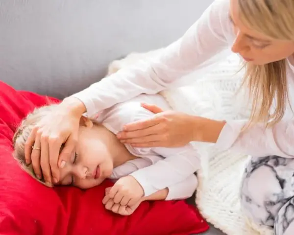 Matka troskliwie opiekuje się dzieckiem, które leży na czerwonej poduszce. Dziecko ma zamknięte oczy, a matka delikatnie dotyka jego czoła, sprawdzając, czy nie ma gorączki. To obrazek pokazujący troskę o dziecko podczas jelitówki.