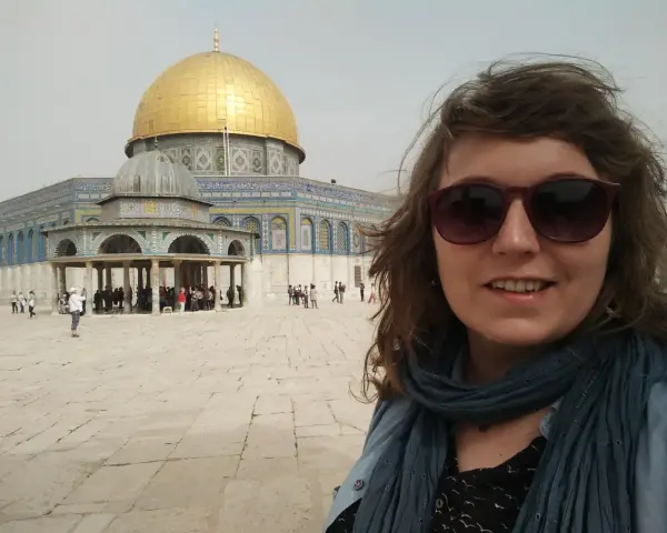 Uśmiechnięta kobieta w okularach przeciwsłonecznych robi selfie na tle Kopuły na Skale. Wokół widać turystów, co sugeruje, że w Jerozolimie jest bezpiecznie.