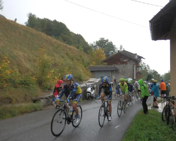 Ciclisti in gara su una strada bagnata, con una **forcella alta** che indica la salita. Il paesaggio è autunnale, con alberi e un pendio erboso.