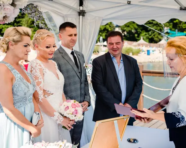 Ceremonia ślubna nad jeziorem, panna młoda w białej sukni, pan młody w garniturze, urzędniczka w przyłbicy.