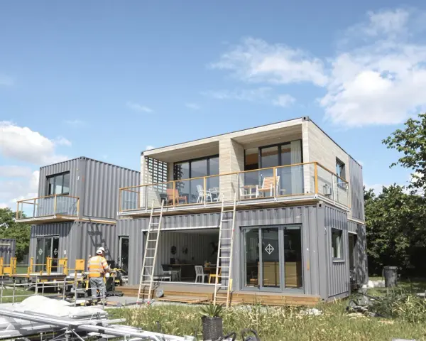 Maison en container prix : construction moderne à deux étages avec balcons, fenêtres panoramiques et un ouvrier en gilet de sécurité.