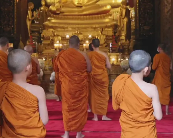 A group of Buddhist monks, another word for ascetics, walk towards a large golden Buddha statue in a temple.
