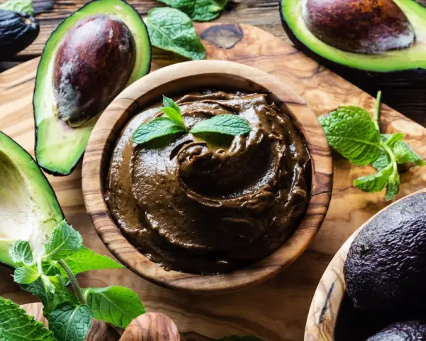 Crema di avocado vellutata in ciotola di legno, circondata da avocado freschi e foglie di menta.