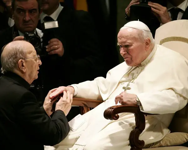 Papież Jan Paweł II w białej szacie, klęczący mężczyzna w czerni. W tle widać fotografów. To obraz duchowej siły, jakiej doświadczają legiony Chrystusa.