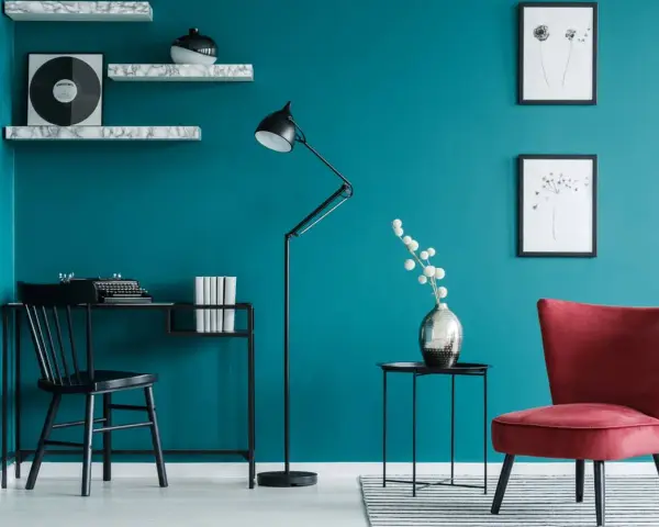 Bureau noir avec machine à écrire, lampe sur pied, fauteuil rouge et étagères sur un mur bleu ou vert canard.