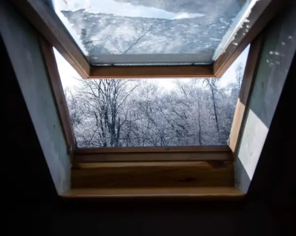 Ein geöffnetes Kellerfenster im Winter gibt den Blick frei auf kahle, schneebedeckte Bäume.