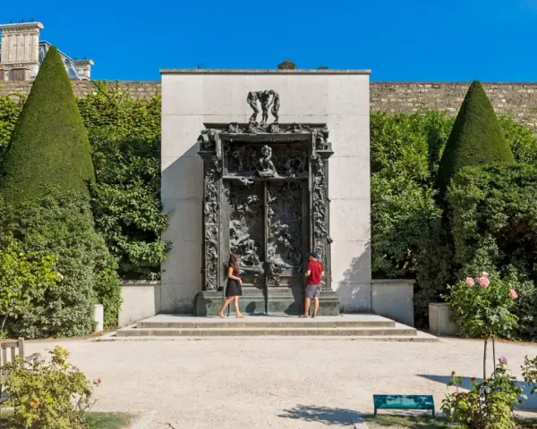 La Porte de l'Enfer, une œuvre monumentale de Rodin, se dresse majestueusement, encadrée par des arbres taillés et un mur de pierre. Deux visiteurs contemplent cette sculpture complexe.
