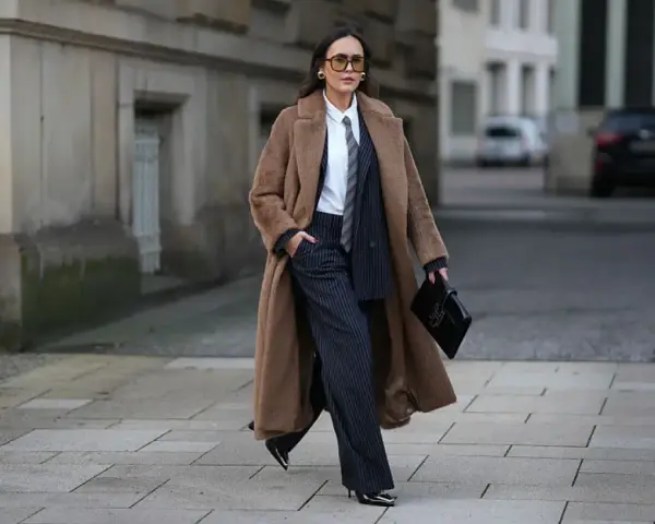 Mujer con abrigo largo marrón, traje de raya diplomática y corbata, luciendo un impecable tomboy style.