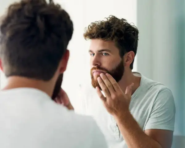 Hombre con barba regia se mira al espejo, acariciando su rostro.