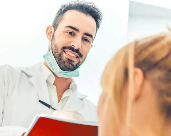 Uśmiechnięty dentysta w białym fartuchu i masce na szyi rozmawia z pacjentką, trzymając notatnik.