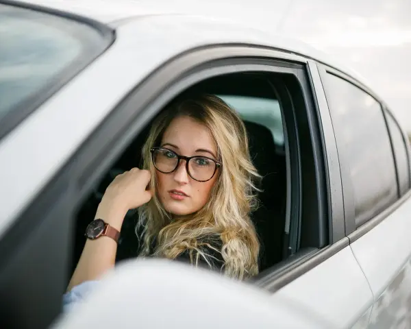 Blondynka w okularach siedzi za kierownicą białego samochodu. Pokazuje, jak nosić okulary, by wyglądać stylowo i pewnie.