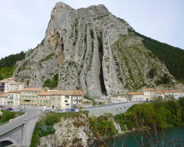 Imponerande klippformation vid Route Napoléon. En by klättrar uppför bergssluttningen, med en bro och en turkos flod nedanför.