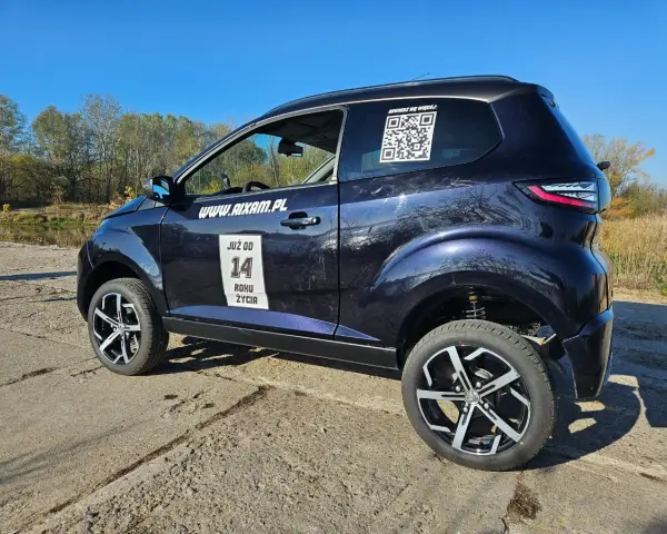 Ciemnoniebieskie Aixam auto z naklejkami "JUŻ OD 14 ROKU ŻYCIA" i kodem QR stoi na tle jesiennego krajobrazu.