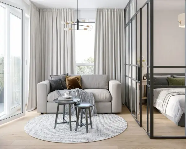 Jasna, nowoczesna kawalerka z wydzieloną sypialnią za szklaną ścianką. W salonie sofa, stoliki i okrągły dywan.