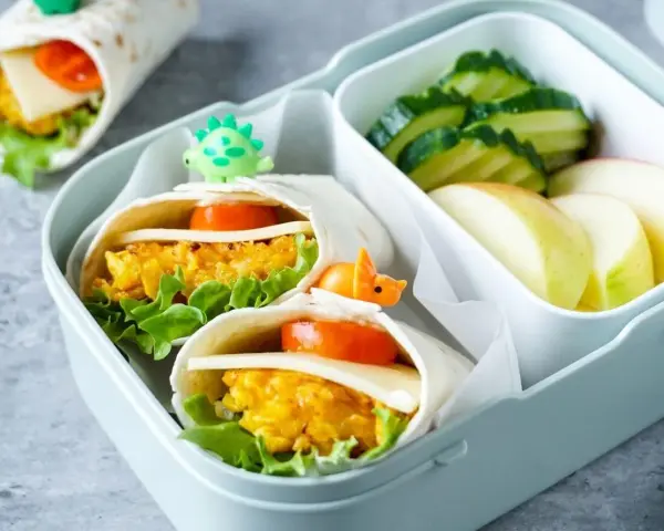 Leckeres Fingerfood für Kinder: Wraps mit Gemüse und Käse, garniert mit kleinen Dino-Spießen. Dazu Gurken und Apfelschnitze.